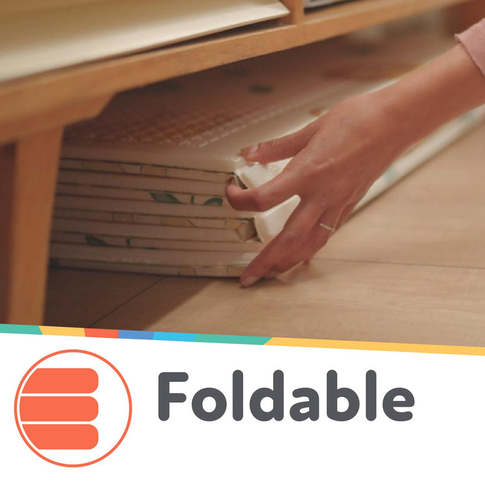 A hand slides the Nuby US Reversible Floor Mat | Graceful Grey under a wooden piece of furniture. "Foldable" and an orange icon with three stacked lines appear at the bottom of the image.