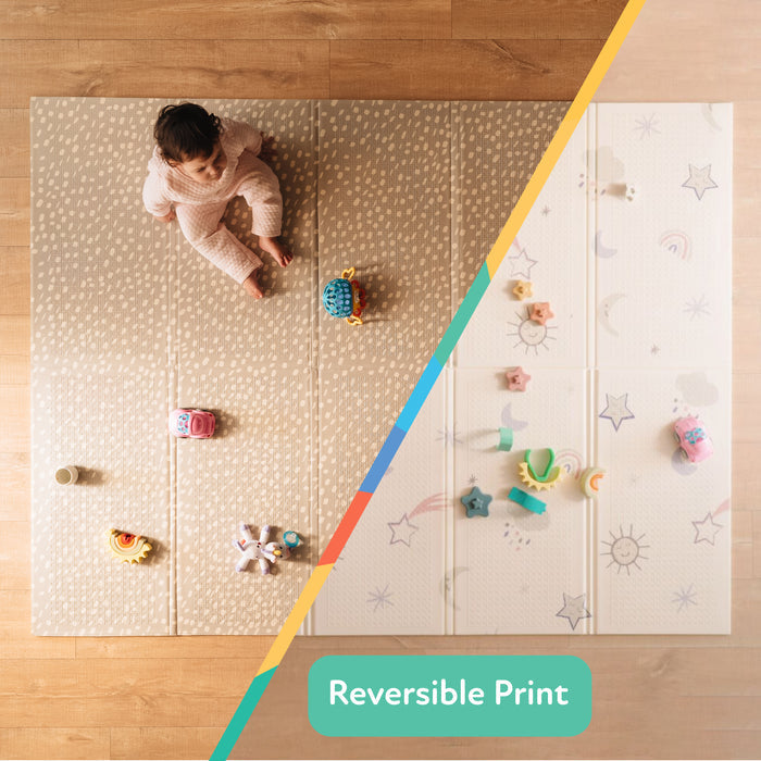 A baby sits on the Nuby US Reversible Floor Mat | Rainbows & Sky. The mat is split diagonally: one side features a subtle beige pattern, the other displays stars, moons, and colorful toys. Text reads “Reversible Print.”.