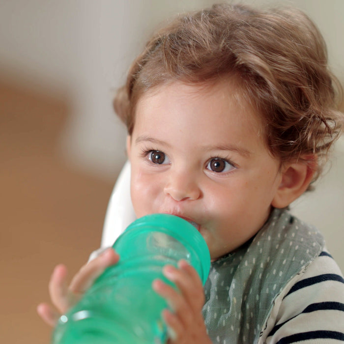 A young child with curly hair drinks from the Nuby Thirsty Kids Sip It Sport Beginner Travel Cup | Rocket, a green, break-resistant cup with a No-Spill spout, while wearing a striped shirt and bib and looking up with wide eyes.